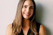 Smiling fair-skinned woman with chest length brown hair leans against a wall