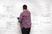 A student wearing a plaid shirt with his back facing the camera, writing on a whiteboard that is covered in notes and diagrams
