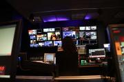 A student in a broadcasting studio, facing multiple monitors that create a large collage of live feeds