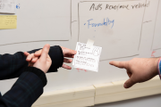 A hand pointing toward a held post-it with a sketch of a user profile in front of a whiteboard