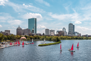 A view of the Boston skyline from the Charles River. Red sailboats are in the water and the sky is bright blue with some clouds.