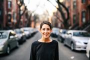 A fair skinned person with pulled back hair smiles in front of a blurry street in the South End