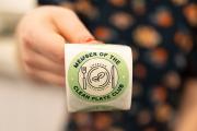 A hand holding a green sticker labeled "Member of the Clean Plate Club," provided by Emerson Sustainability