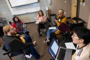 A group of six individuals sitting in a circle in a classroom, engaged in discussion. Some have laptops or notebooks. A whiteboard and projection screen are visible in the background.