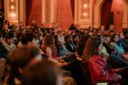 An audience attentively watches a presentation in an ornate, warmly lit theater.