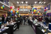 A lively fair takes place in a gymnasium, with colorful balloons and numerous booths staffed by professionals engaging with attendees.