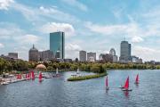 The Boston skyline from the water on a clear summer day; there are small yachts with red sails in the bay.