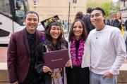 A student smiling while holding her degree cover with family members joined at both sides. They have a floral arrangement as part of their cap and colorful stoles. A person next to her is wearing an orchid lei.