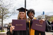 Two graduates wearing regalia and holding their diploma covers up at graduation
