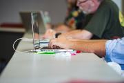 A close-up shows people working at a table with laptops open. Hands are typing on keyboards, with cables and small accessories spread across the surface. The setting feels like a classroom or workshop focused on hands-on computer work.