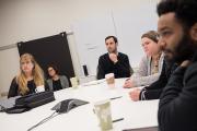 A group of people are seated around a conference table in a meeting room. They appear focused and serious, as if listening or discussing an important topic. Coffee cups, notebooks, and a speakerphone on the table suggest a professional work meeting.