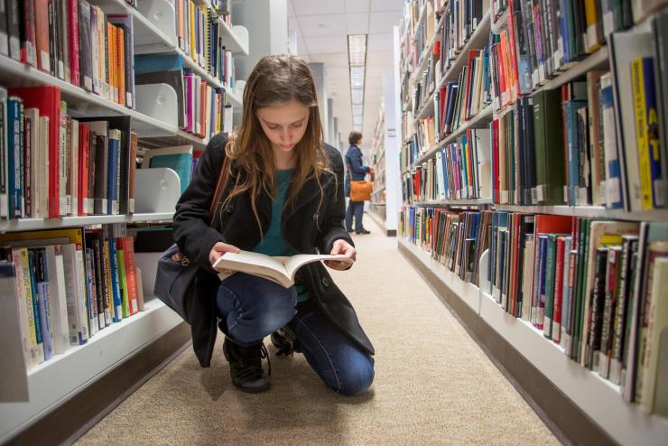 A student kneeling in an aisle at the Iwasaki Library, reading through a book