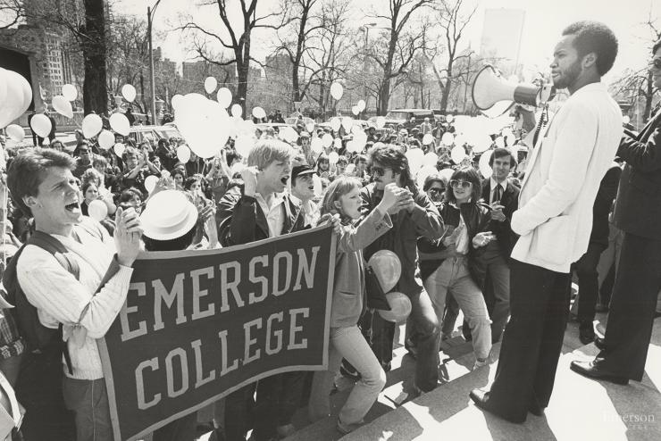 historical image of a crowd of students outside with balloons and Emerson College banner, in front of a stage
