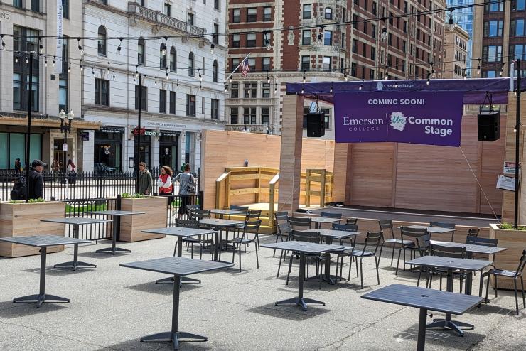 View of the corner of Boylston and Tremont Street, a patio with outdoor tables and chairs are in front of small wooden stage