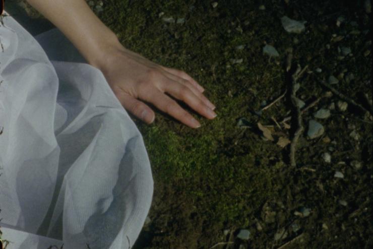 Still from "Reveries Echo of Touch" film, green mossy ground hand touching it with gauzy fabric in frame