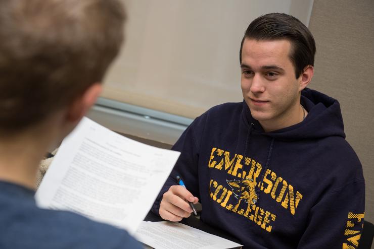 Student in Emerson College sweatshirt holding pen while person in front of them reads off a paper