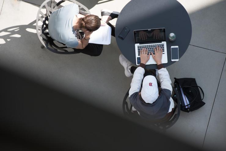 An overhead photo of two students sitting at a table with one on their laptop and the other looking at a paper packet