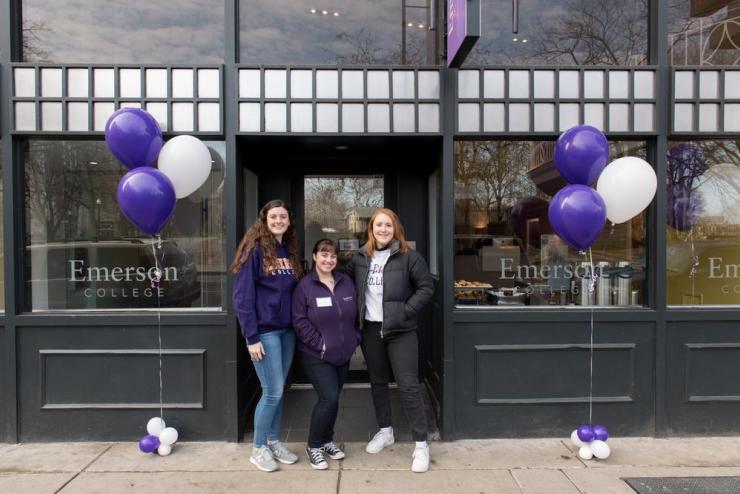 Students wearing Emerson College t-shirts and posing in front of building with purple and white balloons on either side