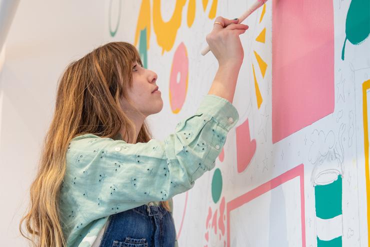 Person with long blonde hair holding paintbrush with pink paint, painting a mural