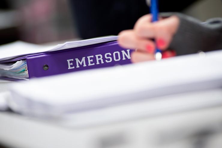 Image of a student's hand writing next to a binder that reads Emerson College