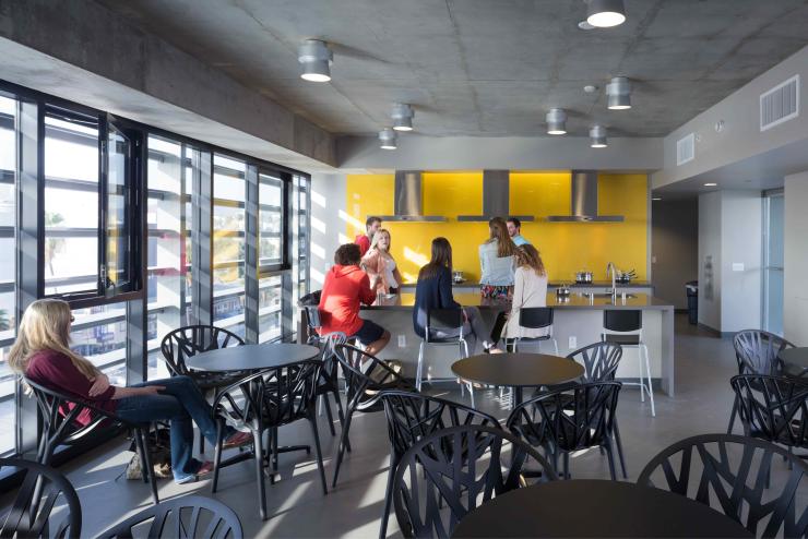 In a sunny dining room with shared kitchen, young people sit at round tables or at the bar