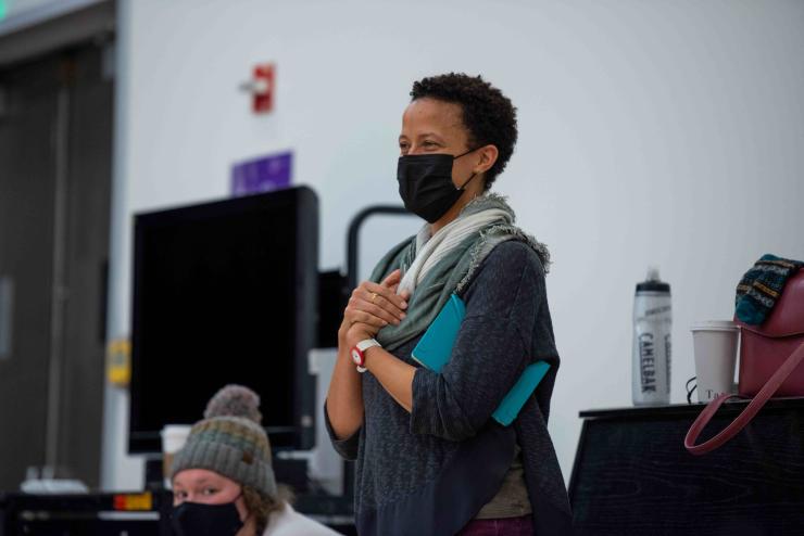 Black woman faculty member with short hair wears a mask and stands smiling, clasping hands