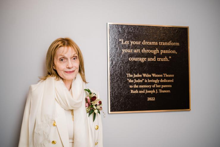 An elder white woman with blonde hair in drapey light-colored outfit stands next to a plaque reading: "Let your dreams transform your art through passion, courage and truth." The Judee Wales Watson Theater "the Judee" is lovingly dedicated to the memory of her parents Ruth and Joseph J. Truncer. 2022