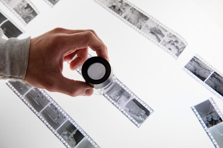 A hand using a magnifying scope to view film strips in a darkroom against a lightboard