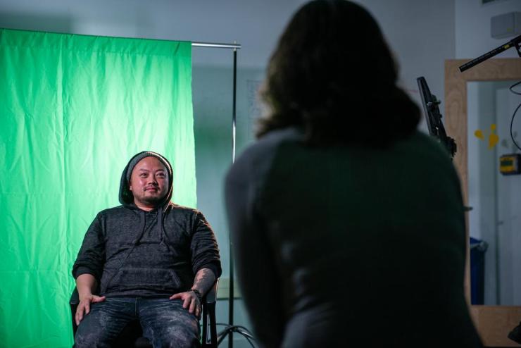 A student sitting casually in front of a curtain green screen, being filmed by another