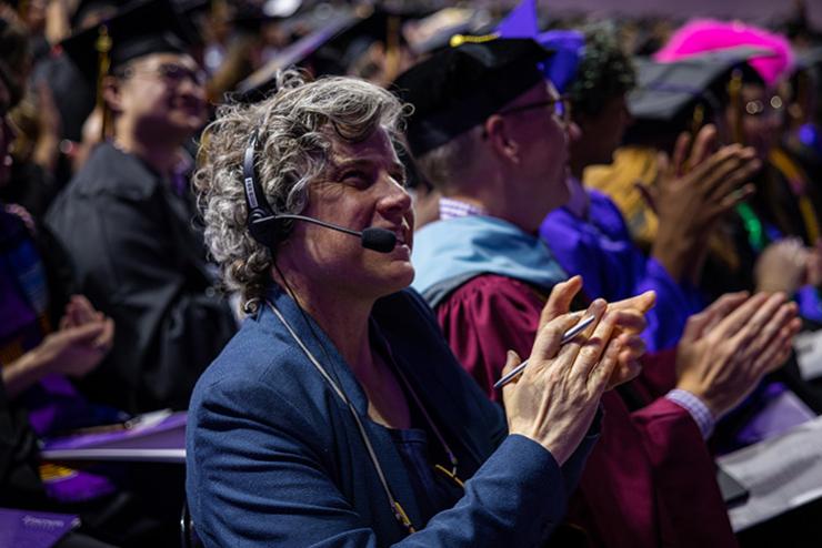 A member of the staff smiles and claps in a room of other people wearing graduation regalia