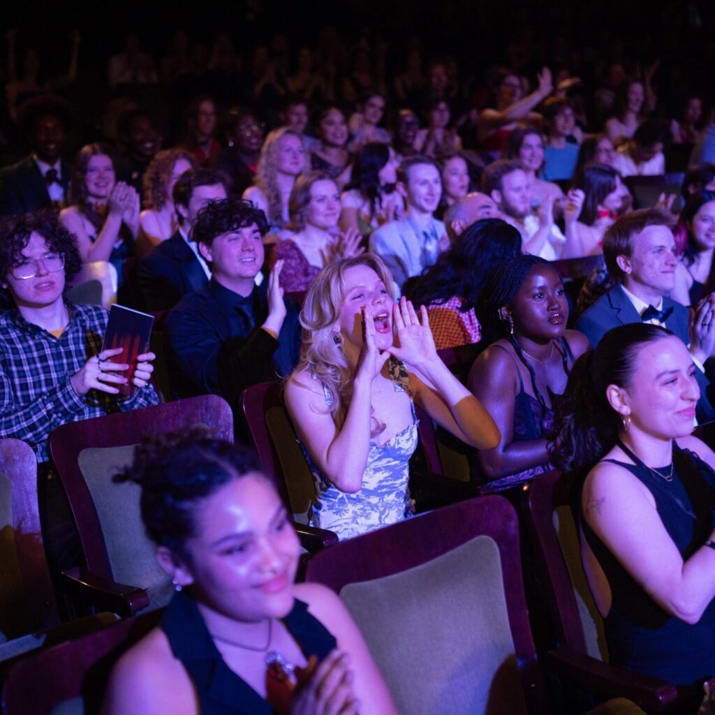 A woman seated in a crowded auditorium frames her hands around her mouth as she cheers.