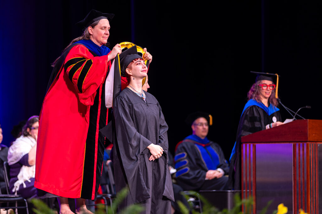 Provost places hood over graduate's head||Graduates wave to processing classmates from seats at the Wang|Man hugs graduate|Graduates cheer|Graduate wearing purple lei poses with friend||Graduate holding diploma