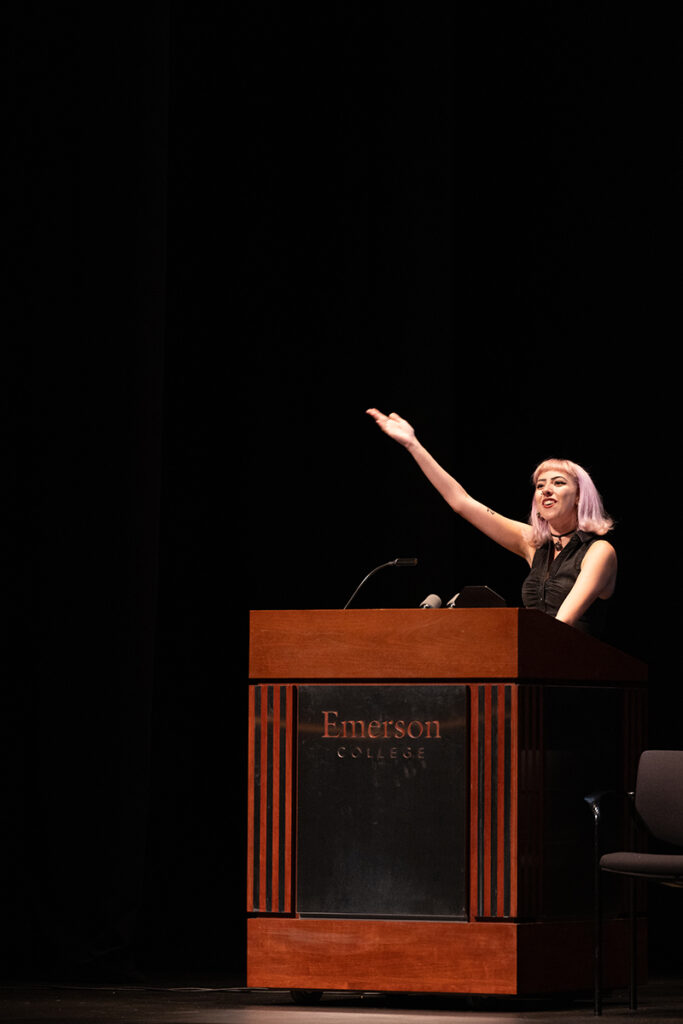 woman in black sleeveless shirt gestures at podium on dark stage