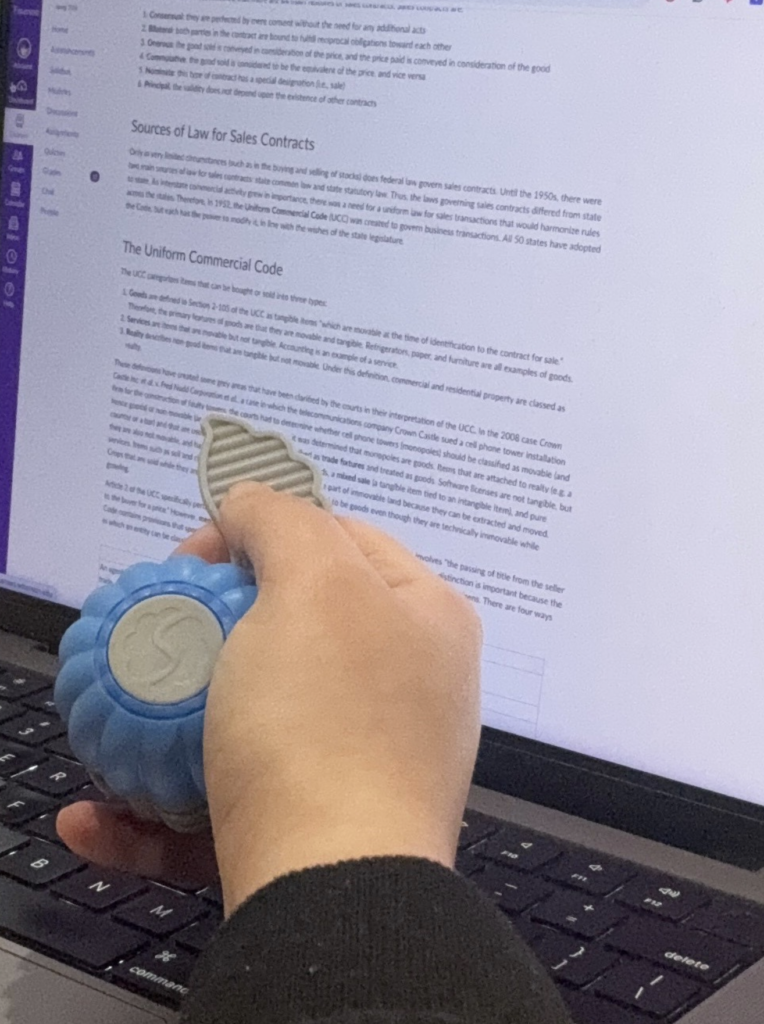 The Sidekick fidget is held in a hand with a computer screen in the background