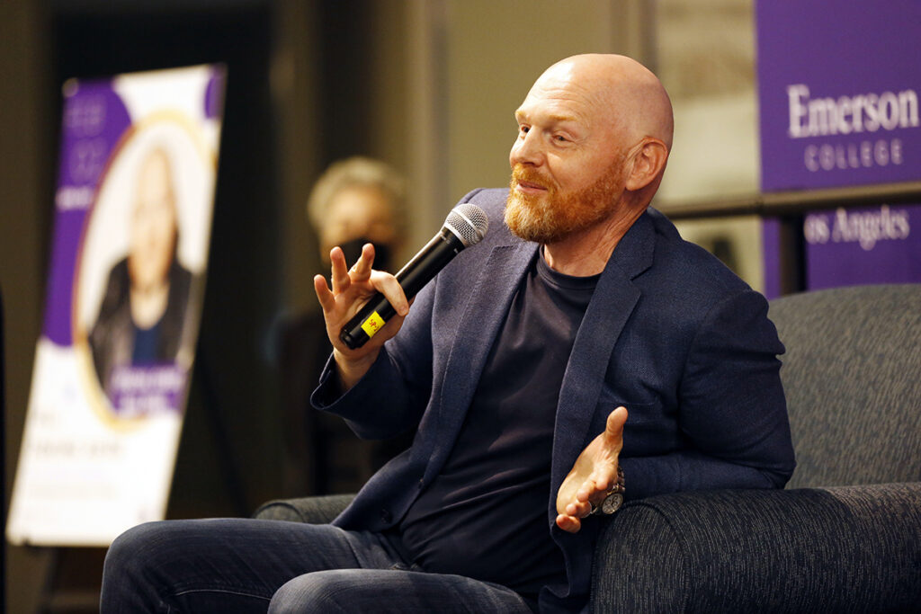Bill Burr in blue blazer leans back in chair, speaks into mic