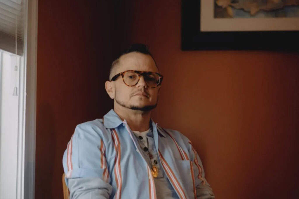 Carl sits against rust colored wall wearing glasses, blue shirt