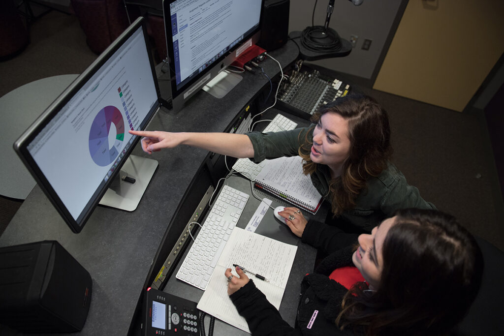 woman points at monitor as another woman takes notes