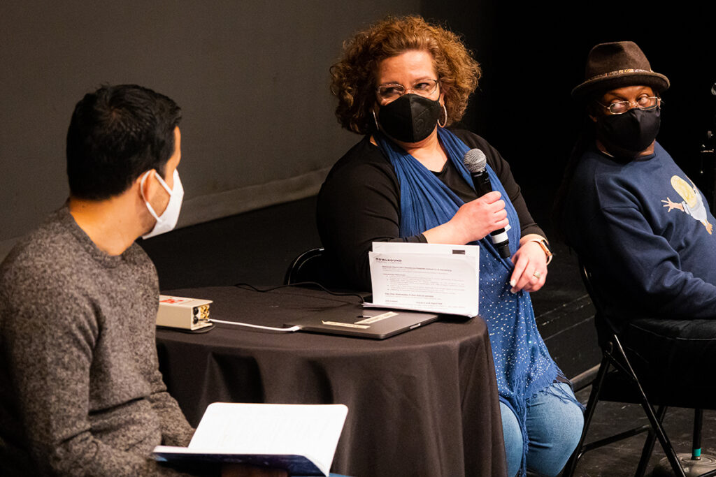 woman holds mic, looks at man holding open book as man on her right looks on