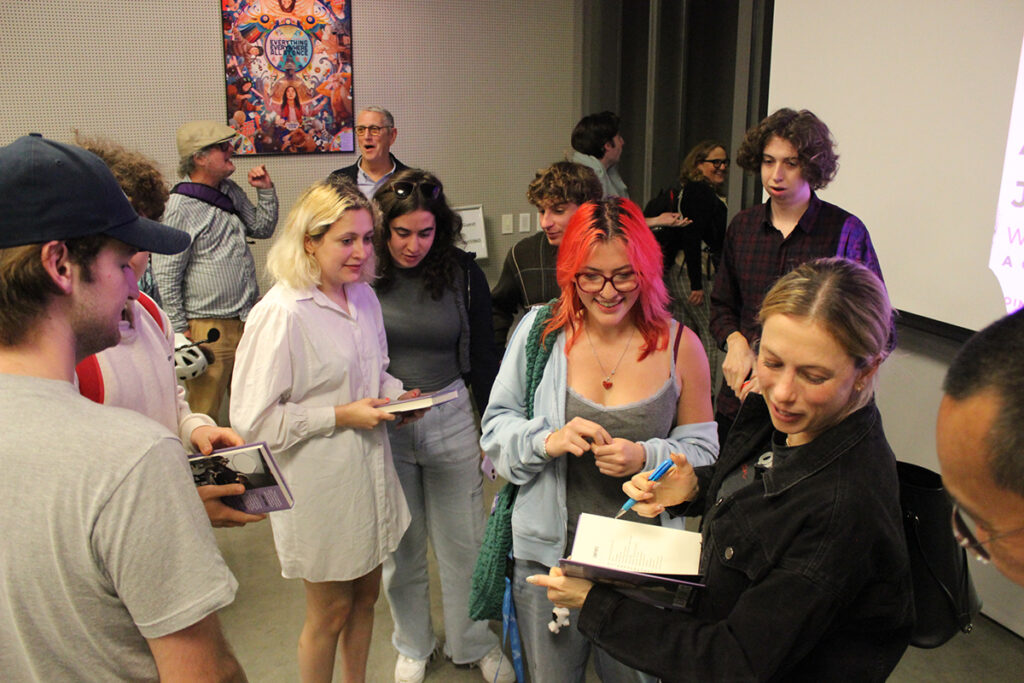 Iliza Shlesinger looks down at something with pen in hand, holding an open book, surrounded by students