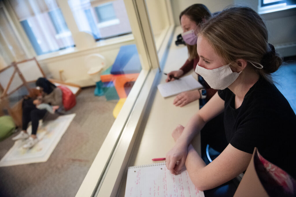 Two clinicians wearing masks through one side of a two-way mirror during a session with a child and parent in a room