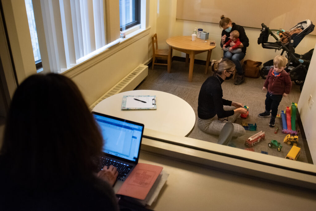 Over the shoulder look behind a two-way mirror while a child works with an adult in a speech therapy room.