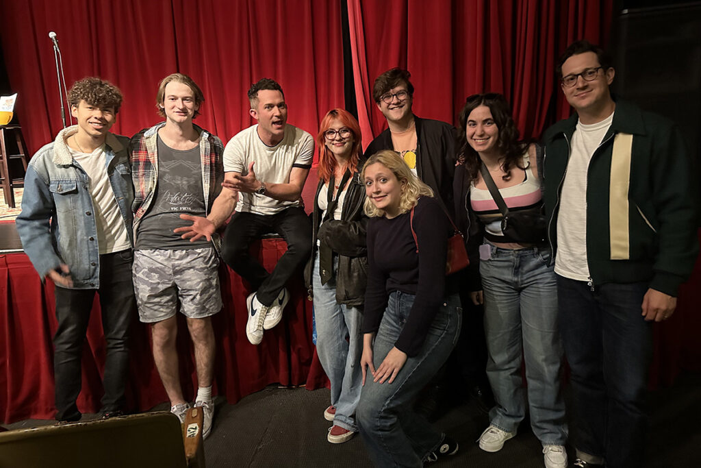 Justin Willman sits on stage surrounded by students