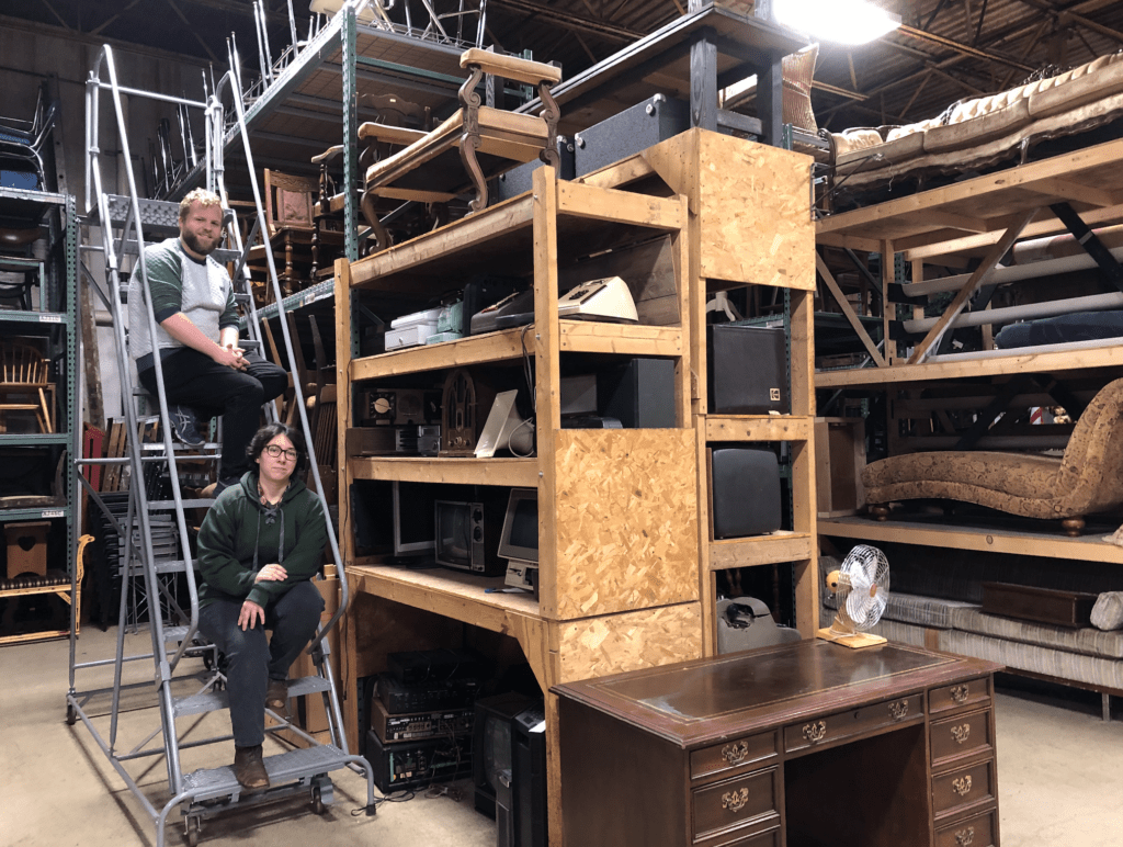 Ryan Bates and Lauren Corcuera sit on a rolling ladder next to wooden shelves packed with stuff