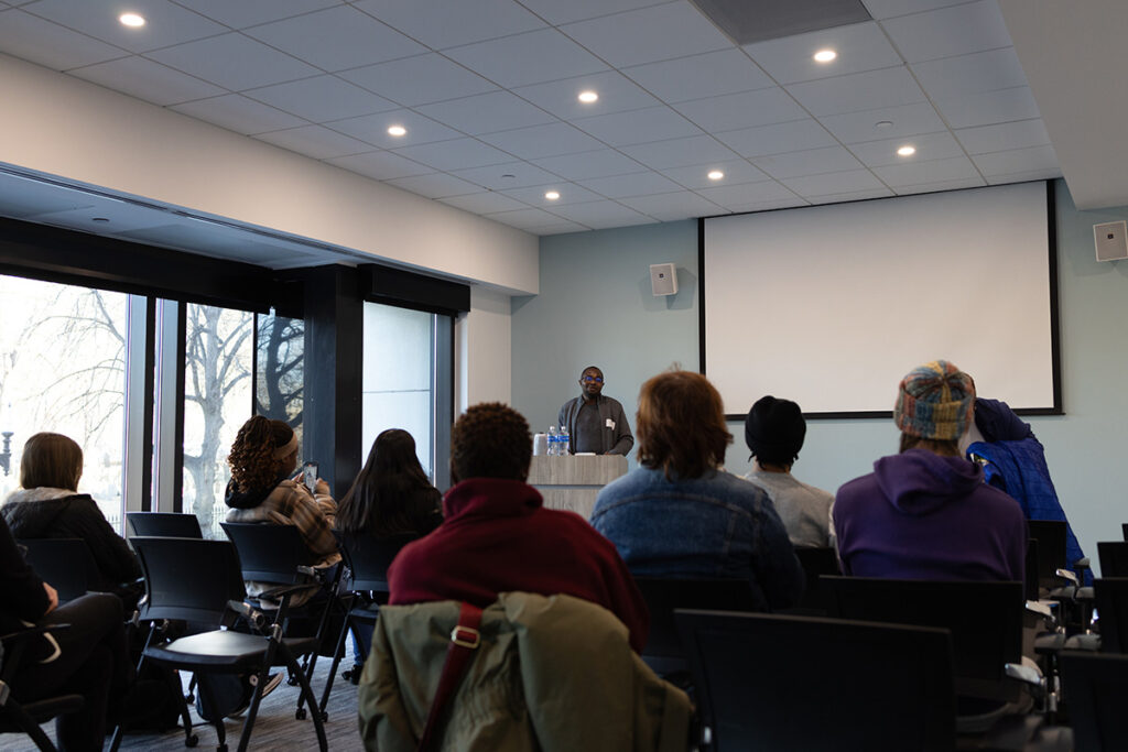 Man speaks at podium as audience listens. Large window is to the left
