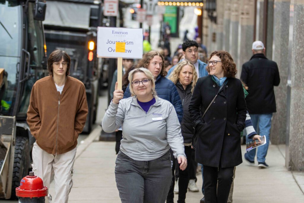 A person walks up the street holding a sign that says they're leading a Journalism Department tour, and there are some people behind them