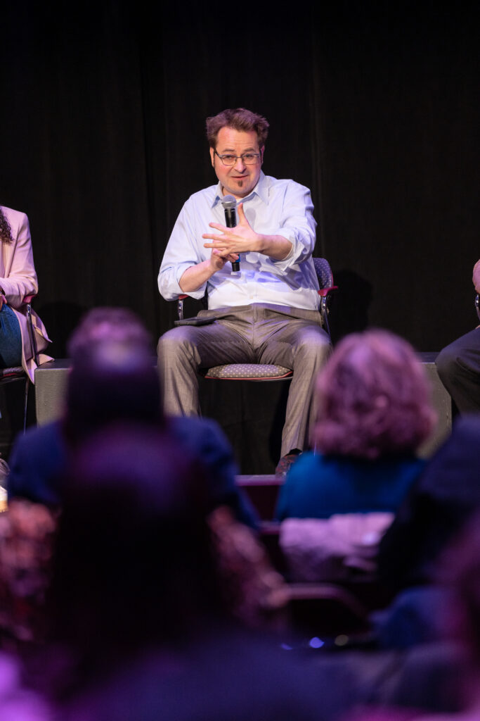 Russell Newman speaks while holding a microphone and sitting in a chair
