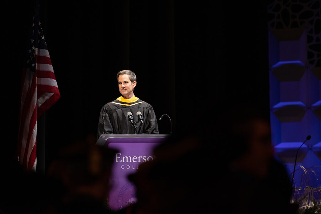 man in black robe stands at purple podium, American flag to his left