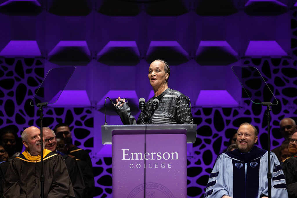woman in black blouse stands at podium as Trustee Chair and President look on and laugh