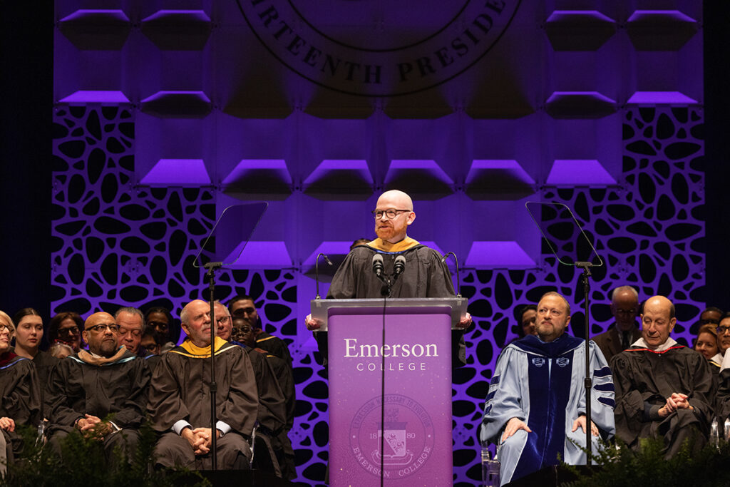Man in black robe stands at podium flanked by trustees and president in regalia