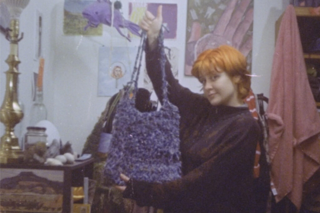 A person holds up a purple bag while looking at the camera